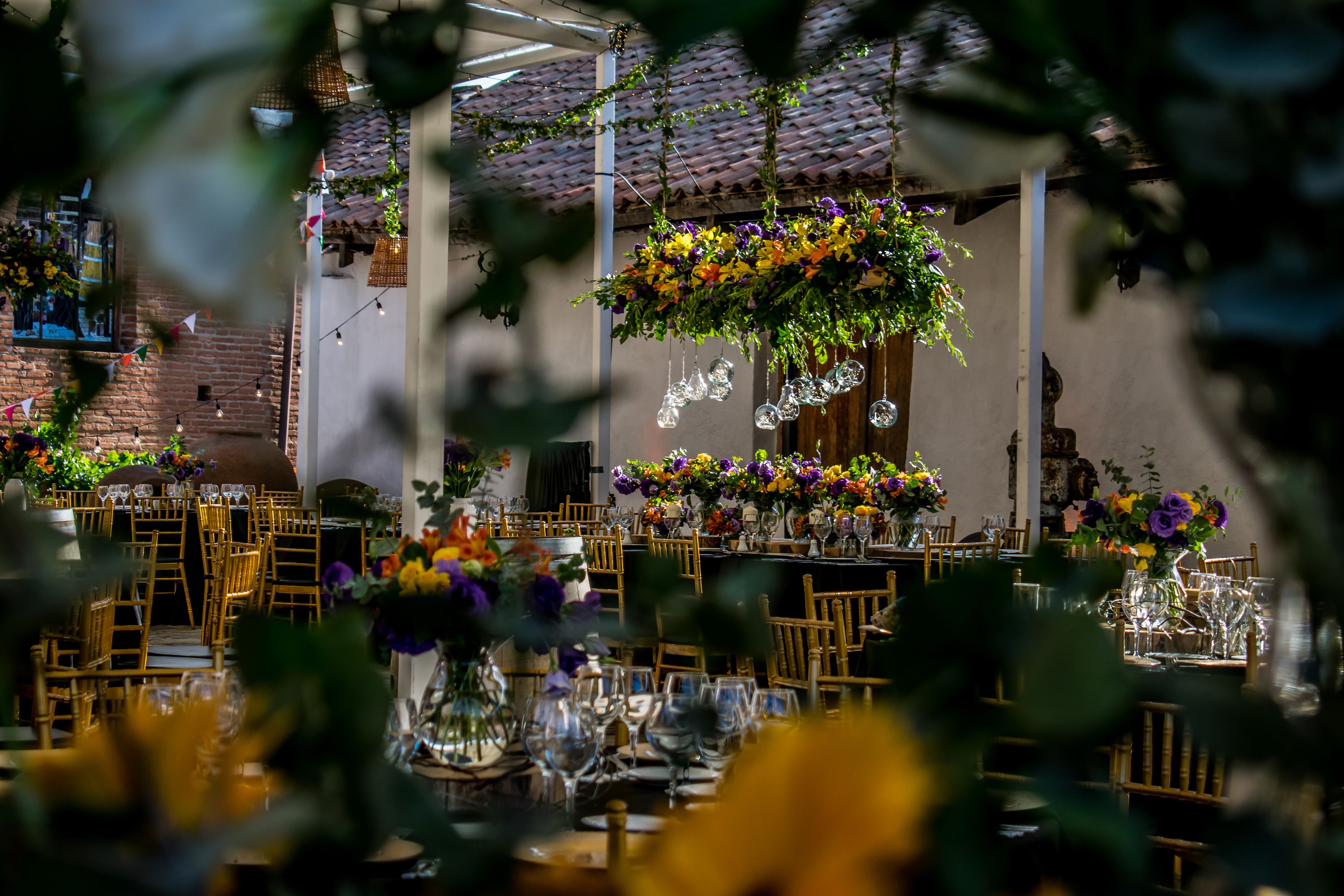 Recepción de matrimonio en un patio con follaje colgante, mesas de madera y centros de mesa florales